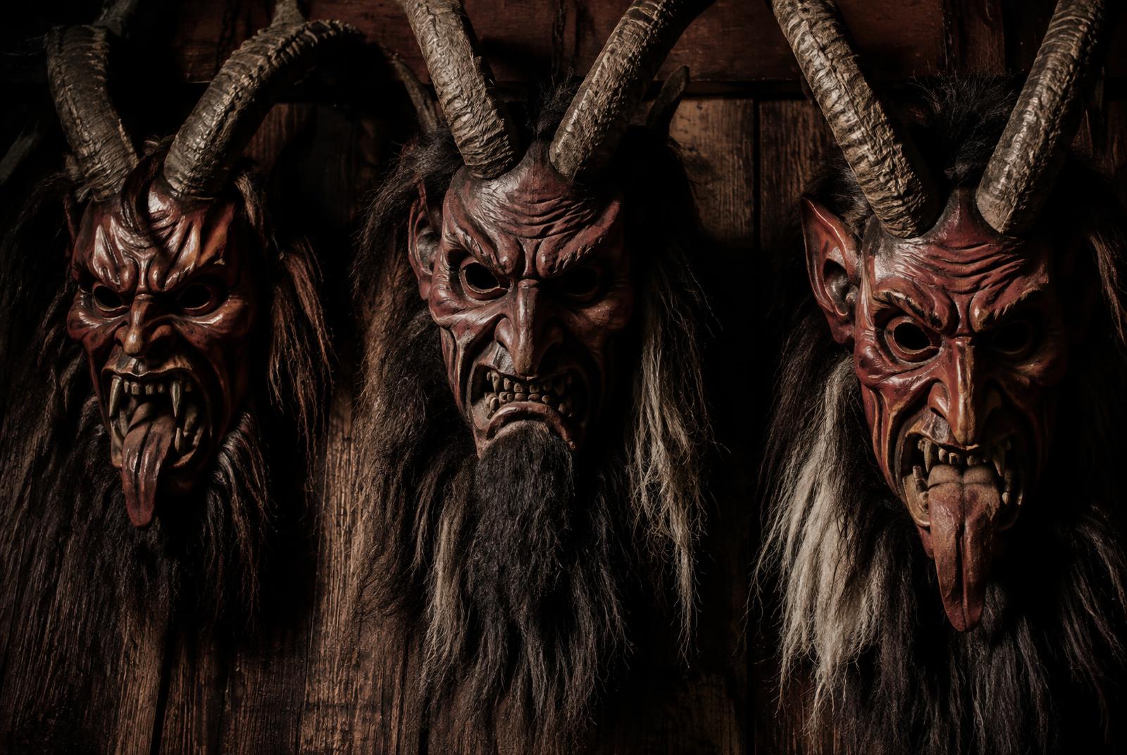 Drei geschnitzte Krampusmasken mit Hörnern und Fell an einer Holzwand (Illustration)